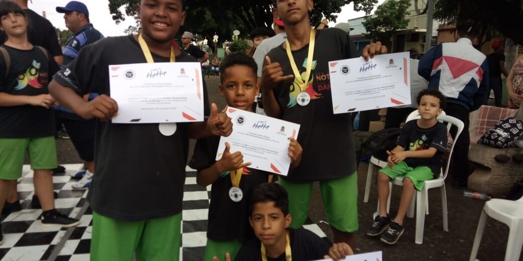 Meninos premiados em evento. Quatro meninos sorridentes mostram certificados e medalhas em um evento ao ar livre, enquanto uma criança sentada observa ao fundo.