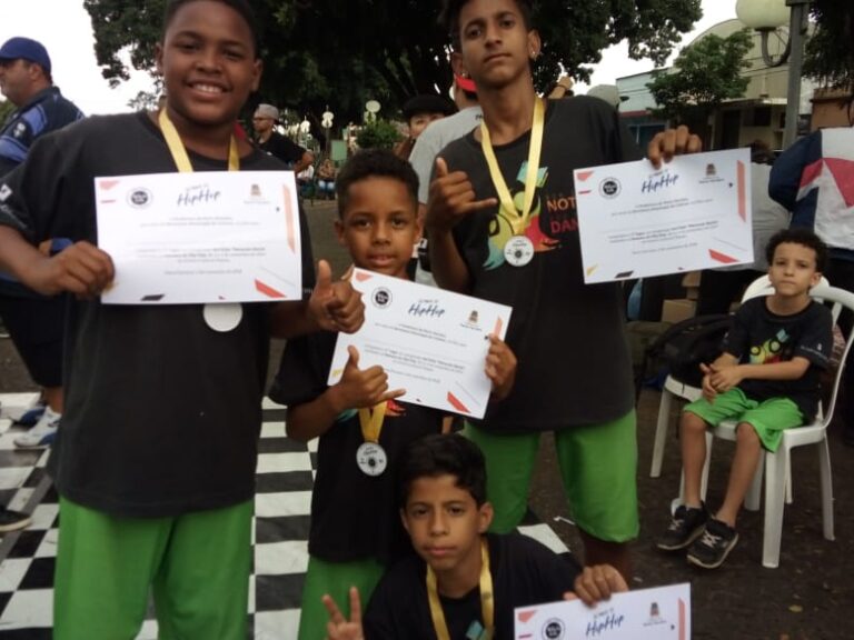 Meninos premiados em evento. Quatro meninos sorridentes mostram certificados e medalhas em um evento ao ar livre, enquanto uma criança sentada observa ao fundo.