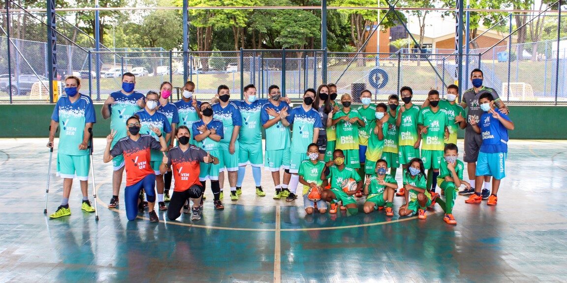 Grupo de crianças e adultos uniformizados posa para foto em quadra. Grande grupo de crianças e adultos com uniformes azuis e verdes, e máscaras, posando em uma quadra externa.
