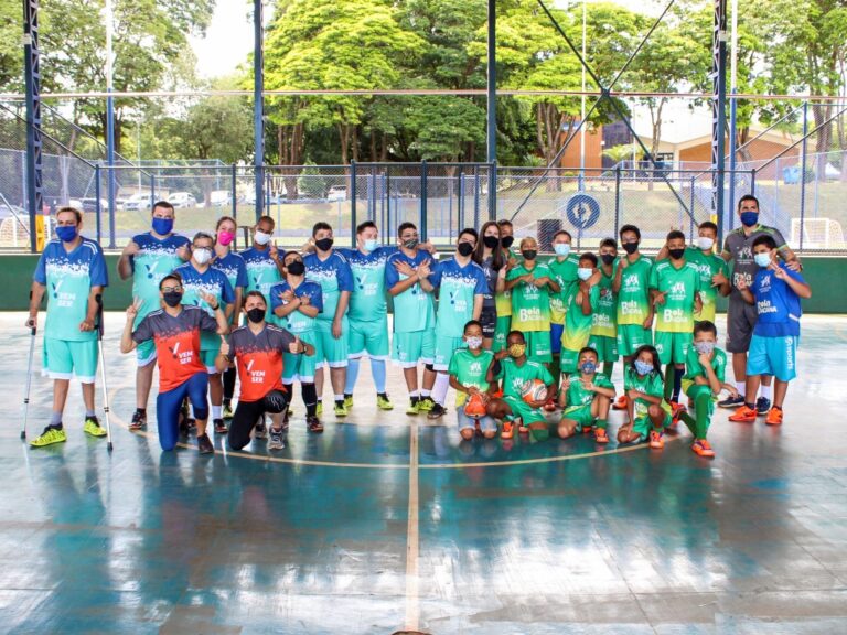 Grupo de crianças e adultos uniformizados posa para foto em quadra. Grande grupo de crianças e adultos com uniformes azuis e verdes, e máscaras, posando em uma quadra externa.