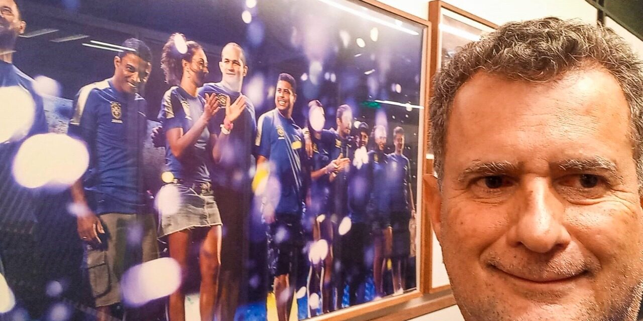 Selfie ao lado de pôster de atletas comemorando. Selfie de um homem sorridente ao lado de um pôster emoldurado, com atletas em camisas azuis comemorando.