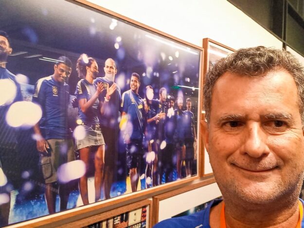 Selfie ao lado de pôster de atletas comemorando. Selfie de um homem sorridente ao lado de um pôster emoldurado, com atletas em camisas azuis comemorando.