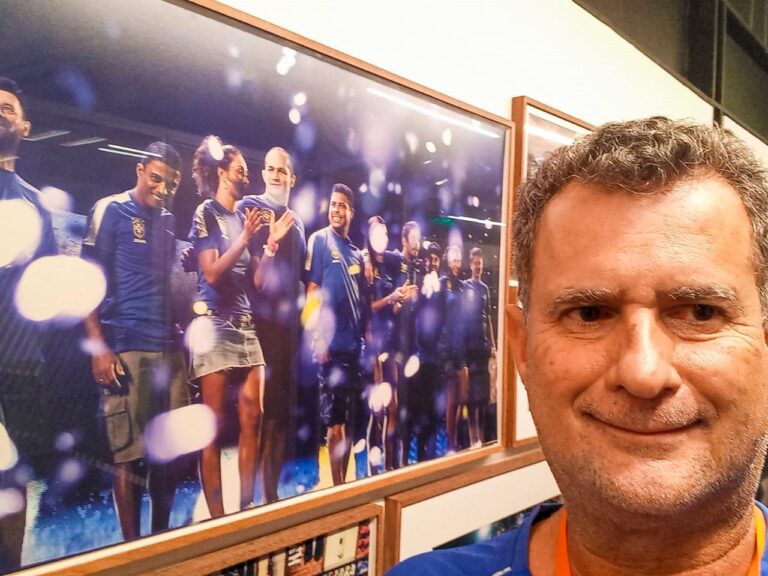 Selfie ao lado de pôster de atletas comemorando. Selfie de um homem sorridente ao lado de um pôster emoldurado, com atletas em camisas azuis comemorando.