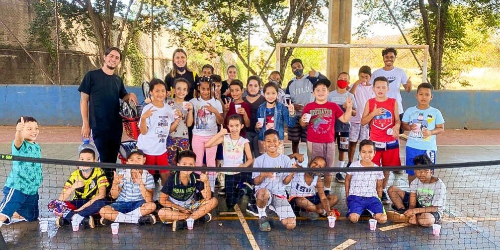 Crianças e instrutores em quadra esportiva. Grupo de cerca de 25 crianças e três adultos em quadra esportiva, posando para foto em frente a uma rede baixa de tênis.