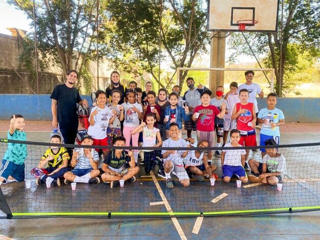 Crianças e instrutores em quadra esportiva. Grupo de cerca de 25 crianças e três adultos em quadra esportiva, posando para foto em frente a uma rede baixa de tênis.