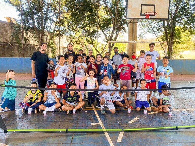 Crianças e instrutores em quadra esportiva. Grupo de cerca de 25 crianças e três adultos em quadra esportiva, posando para foto em frente a uma rede baixa de tênis.