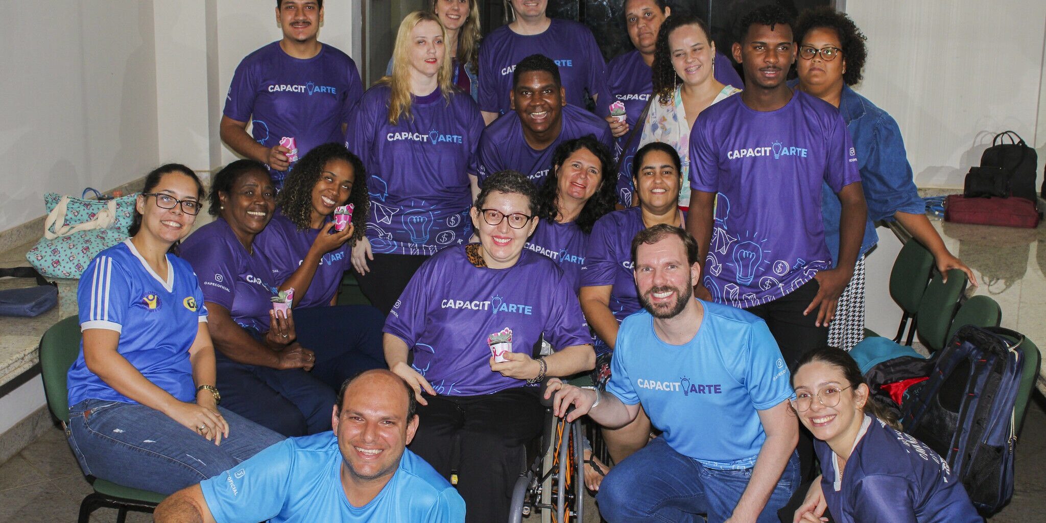 Grupo diverso de participantes do projeto. Grupo diverso de cerca de 18 pessoas, a maioria com camisetas CapacitArte roxas e azuis, posando juntas para foto.