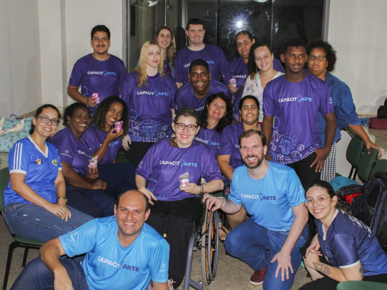 Grupo diverso de participantes do projeto. Grupo diverso de cerca de 18 pessoas, a maioria com camisetas CapacitArte roxas e azuis, posando juntas para foto.