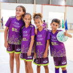 Quatro crianças, três meninas e um menino, vestindo uniformes de futebol roxos com detalhes verdes, posam juntas. O menino segura uma bola de futebol e faz um sinal de "joinha" com o polegar.