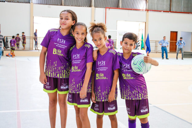 Quatro crianças, três meninas e um menino, vestindo uniformes de futebol roxos com detalhes verdes, posam juntas. O menino segura uma bola de futebol e faz um sinal de "joinha" com o polegar.