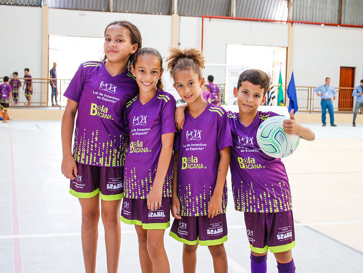 Quatro crianças, três meninas e um menino, vestindo uniformes de futebol roxos com detalhes verdes, posam juntas. O menino segura uma bola de futebol e faz um sinal de "joinha" com o polegar.