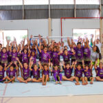 Grande grupo de crianças e adolescentes, usando uniformes roxos, posa em quadra com as mãos levantadas, empolgados com o projeto esportivo.