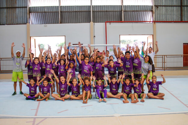 Grande grupo de crianças e adolescentes, usando uniformes roxos, posa em quadra com as mãos levantadas, empolgados com o projeto esportivo.