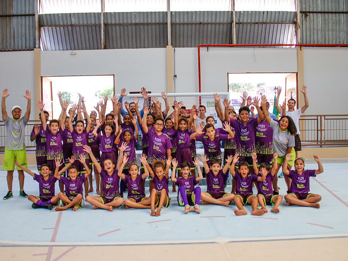 Grande grupo de crianças e adolescentes, usando uniformes roxos, posa em quadra com as mãos levantadas, empolgados com o projeto esportivo.