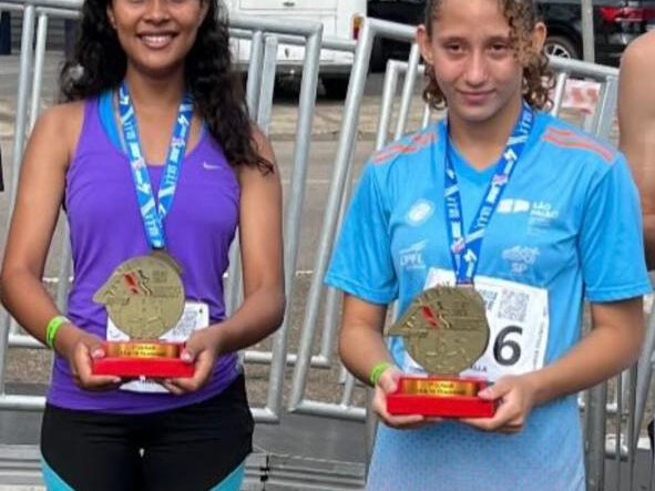 Alunas do vamos pra cima laranja vencem corrida de rua em Sumaré. Duas jovens atletas sorridentes, usando uniformes esportivos, exibem medalhas de ouro e troféus em uma competição.