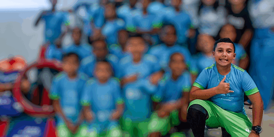 Aluno do projeto bola bacana 11 Garoto sorridente em primeiro plano, agachado e fazendo um polegar para cima, com um grupo de crianças uniformizadas desfocado ao fundo.