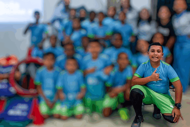Aluno do projeto bola bacana 11 Garoto sorridente em primeiro plano, agachado e fazendo um polegar para cima, com um grupo de crianças uniformizadas desfocado ao fundo.