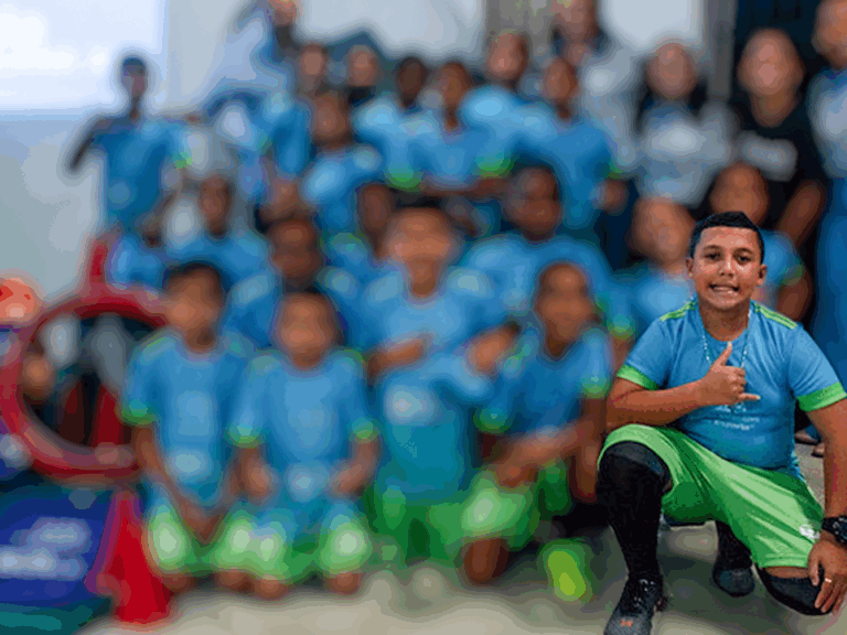 Aluno do projeto bola bacana 11 Garoto sorridente em primeiro plano, agachado e fazendo um polegar para cima, com um grupo de crianças uniformizadas desfocado ao fundo.