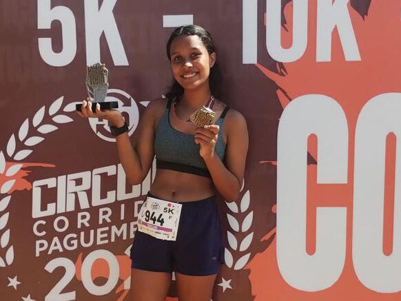 Aluna do vamos pra cima laranja vence terceira corrida de rua. Jovem corredora sorridente, segurando um troféu e uma medalha após a corrida, em frente ao banner do evento.