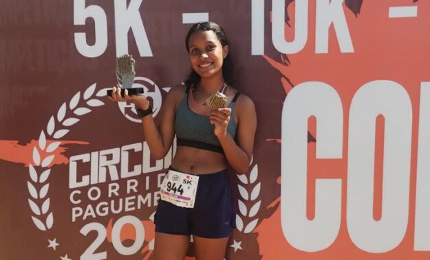 Aluna do vamos pra cima laranja vence terceira corrida de rua. Jovem corredora sorridente, segurando um troféu e uma medalha após a corrida, em frente ao banner do evento.