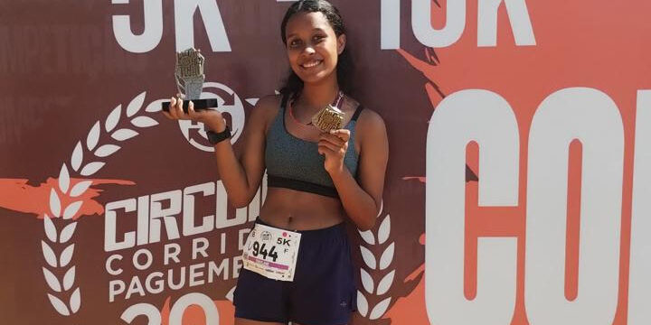 Aluna do vamos pra cima laranja vence terceira corrida de rua. Jovem corredora sorridente, segurando um troféu e uma medalha após a corrida, em frente ao banner do evento.