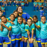 Oito crianças e uma professora posam sorridentes em quadra coberta, com uniformes azul e amarelo, transmitindo alegria e união.