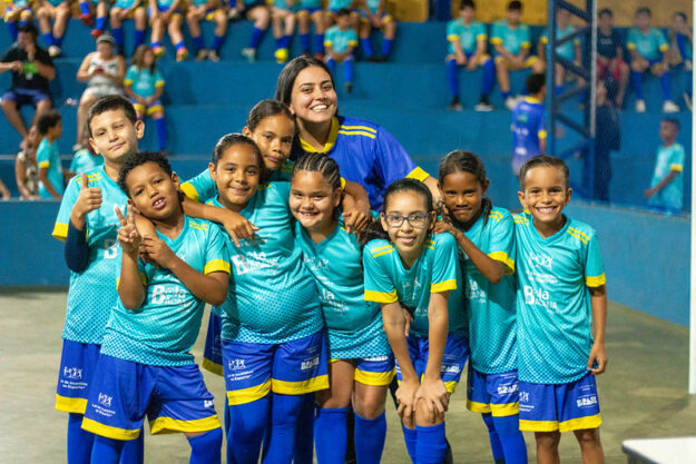 Oito crianças e uma professora posam sorridentes em quadra coberta, com uniformes azul e amarelo, transmitindo alegria e união.