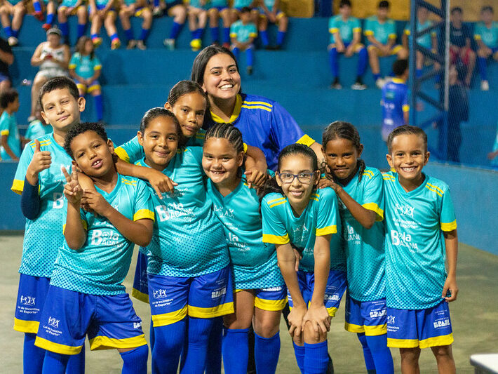 Oito crianças e uma professora posam sorridentes em quadra coberta, com uniformes azul e amarelo, transmitindo alegria e união.