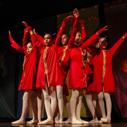 Grupo de meninas no palco executando coreografia. Elas vestem túnicas vermelhas com detalhes dourados e meias-calças brancas, braços levantados.