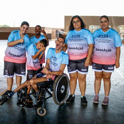 Participantes de projeto esportivo inclusivo comemoram em grupo. Um jovem cadeirante sorri no centro da foto.