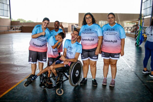 Participantes de projeto esportivo inclusivo comemoram em grupo. Um jovem cadeirante sorri no centro da foto.