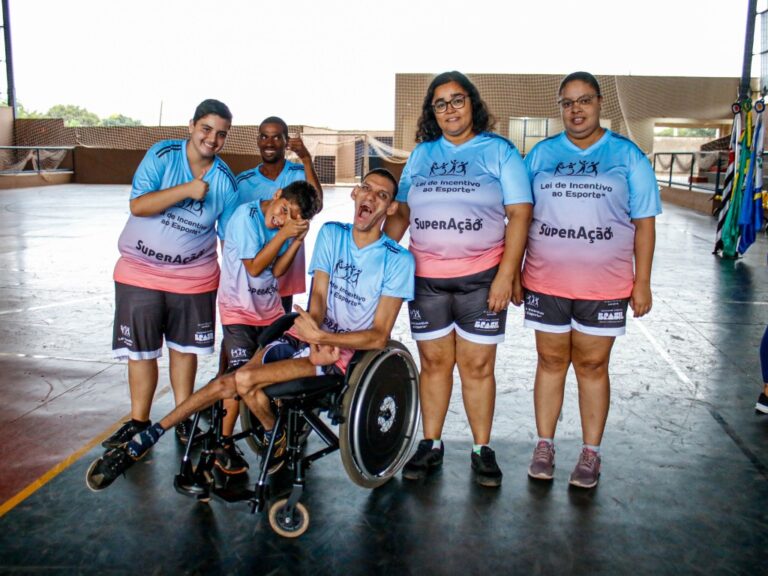 Participantes de projeto esportivo inclusivo comemoram em grupo. Um jovem cadeirante sorri no centro da foto.