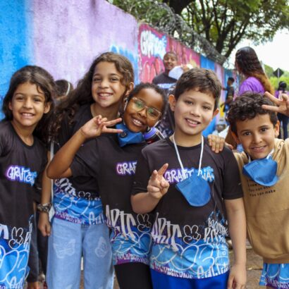 Cinco crianças sorrindo posam em frente a um muro colorido com grafites. Quatro vestem camisetas “GRAFITARTE” e fazem sinal de paz.