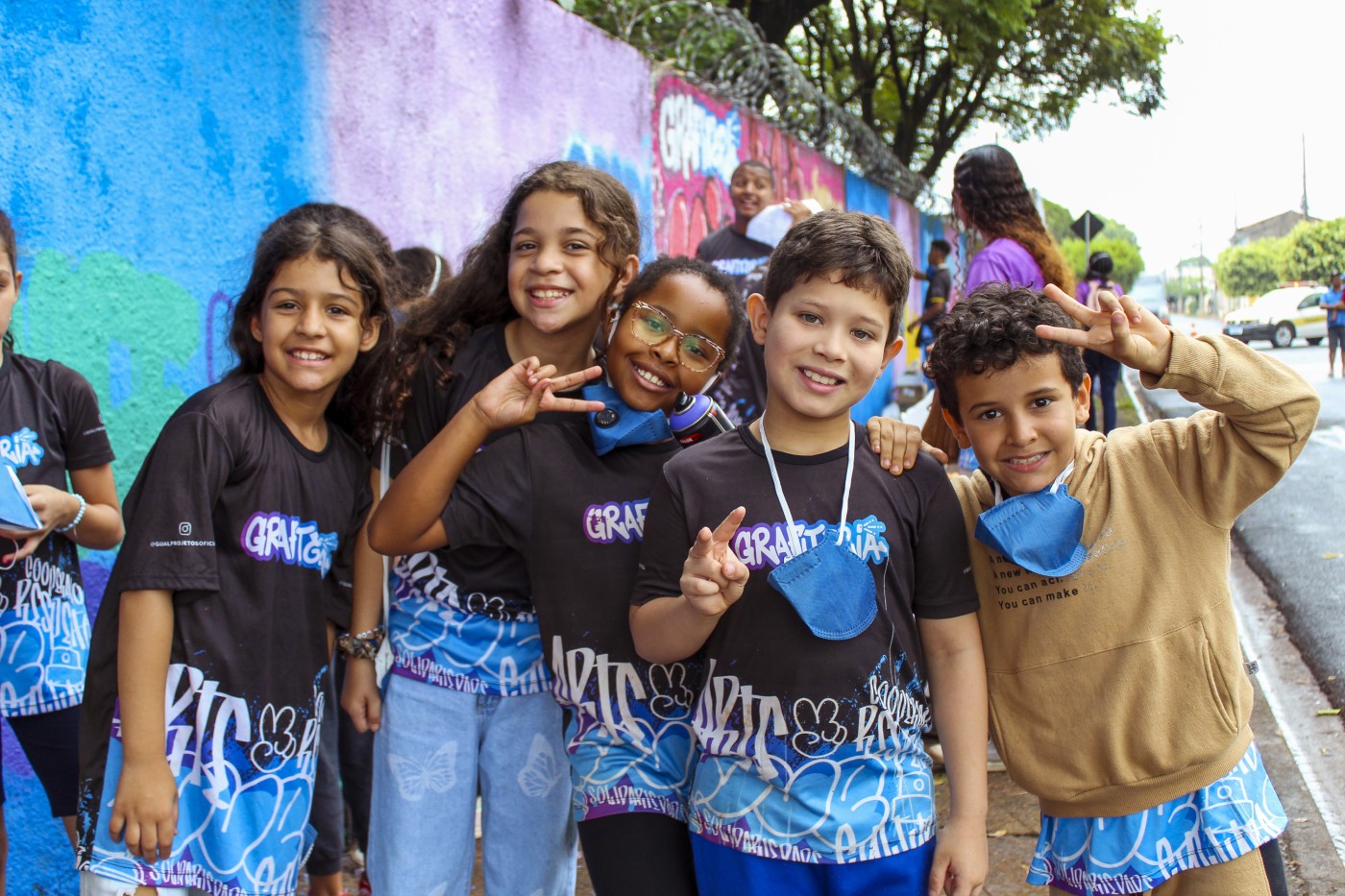 Cinco crianças sorrindo posam em frente a um muro colorido com grafites. Quatro vestem camisetas “GRAFITARTE” e fazem sinal de paz.