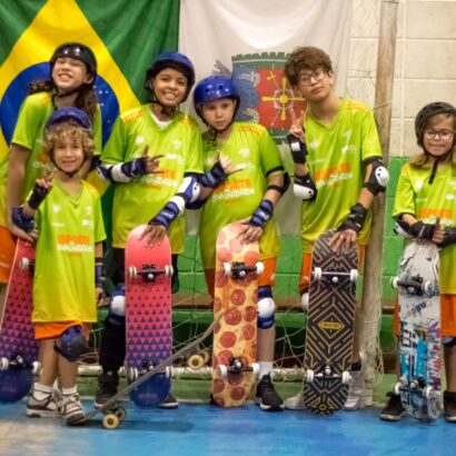 Seis crianças em fila em ginásio, com skates coloridos, capacetes e camisetas verdes. Sorriem e fazem gestos de “V”, transmitindo alegria e união.
