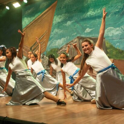 Grupo de jovens dançarinas em palco, realizando coreografia. Vestem roupas brancas e saias cinzas com faixas azuis. Um fundo pintado de barco aparece.