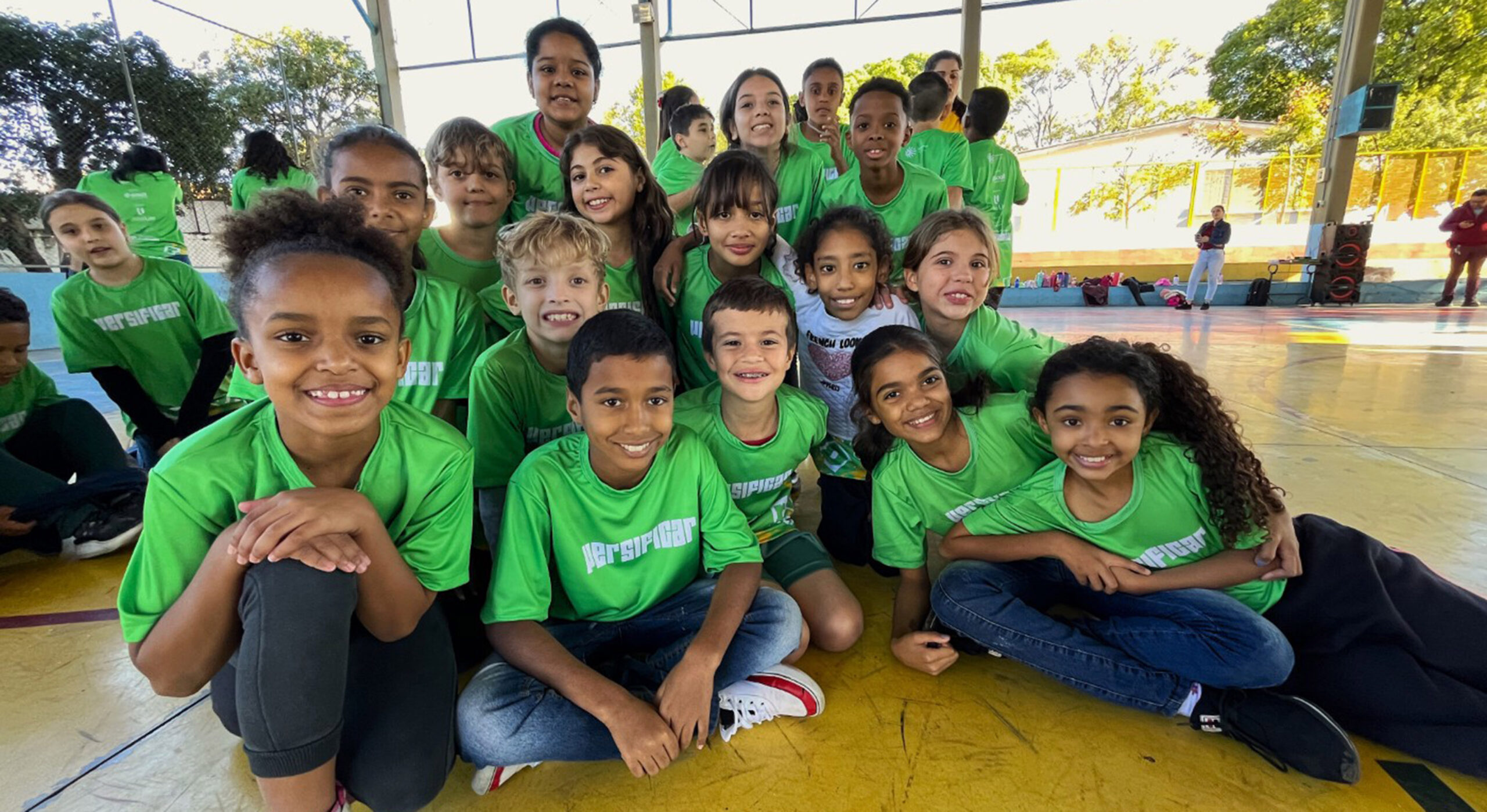 Grupo de crianças sorridentes, usando camisetas verde-limão, posando em quadra de esportes ao ar livre. Algumas estão sentadas na frente.