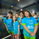 Grupo de cinco crianças sorrindo e posando, em quadra coberta. Todos vestem uniformes de esporte em tons de azul e verde-limão.