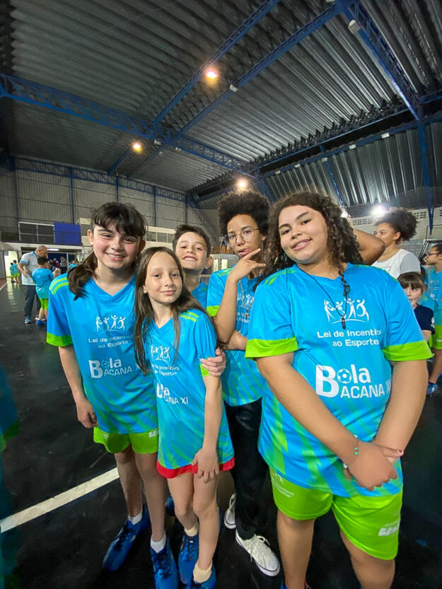 Grupo de cinco crianças sorrindo e posando, em quadra coberta. Todos vestem uniformes de esporte em tons de azul e verde-limão.