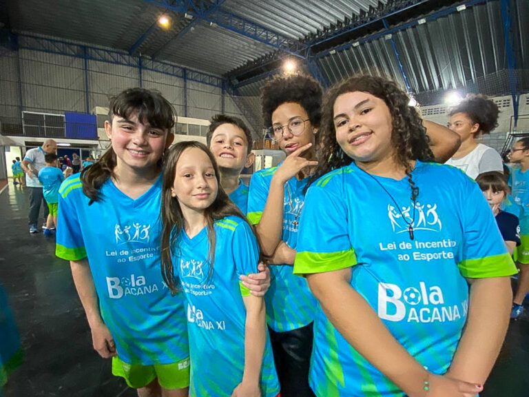 Grupo de cinco crianças sorrindo e posando, em quadra coberta. Todos vestem uniformes de esporte em tons de azul e verde-limão.