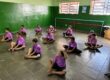 Grupo de doze meninos em camisetas roxas sentados no chão de um estúdio de dança na aula de balé.