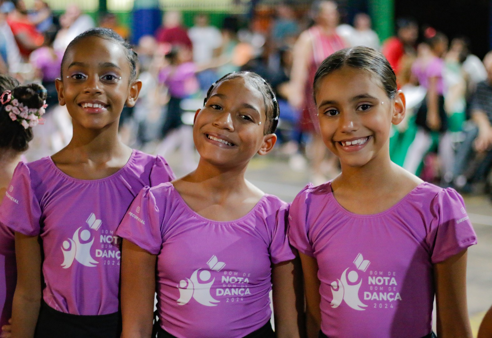 Três meninas sorridentes, usando blusas de dança roxas com estampa branca, em um evento noturno.